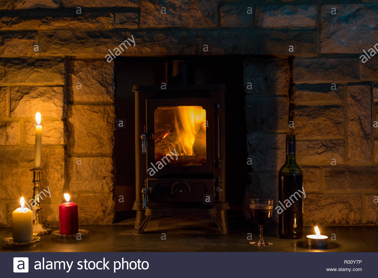 hygge concept a log burner candles and a wine bottle and glass in a stone fireplace R00Y7P